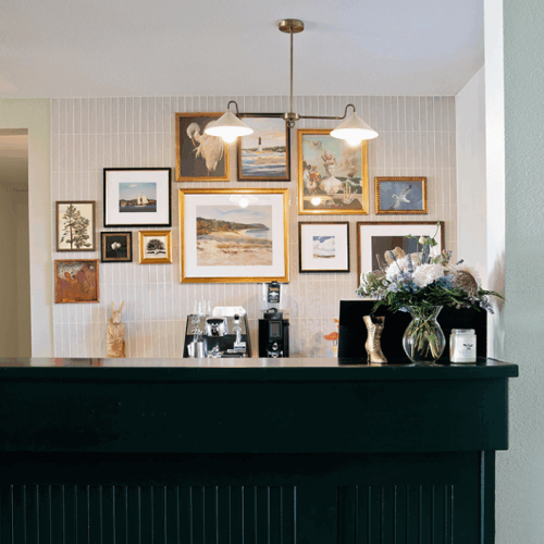 A dark-colored counter with a coffee machine, framed art on the wall, hanging lights, and a bouquet in a vase.