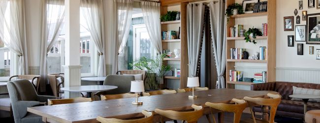A bright, cozy dining space with a long wooden table and mismatched chairs, bookshelves, plants, and a soft, airy vibe with draped windows.