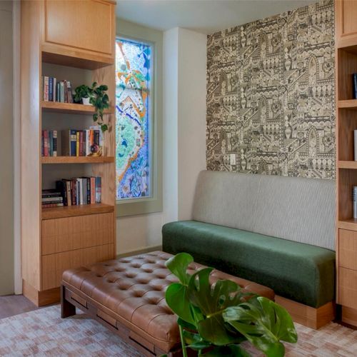 Cozy reading nook with built-in wooden shelves, a green cushioned bench, tufted ottoman, leafy plant, and a patterned wall panel by a window.
