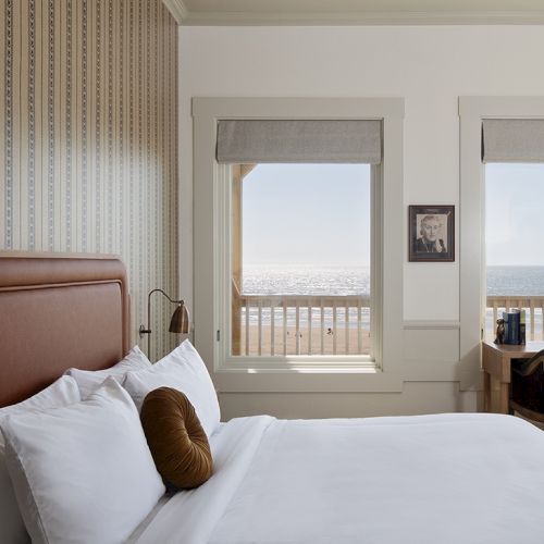 A bright hotel room with a large bed, white bedding, a brown headboard, and a desk by the window overlooking the ocean, sunny and serene.