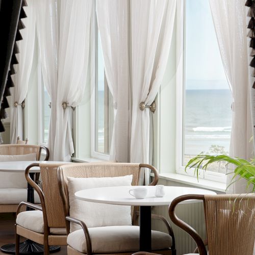 A bright seaside cafe with white draped curtains, cozy round table, and cream chairs by large windows overlooking the ocean breeze.