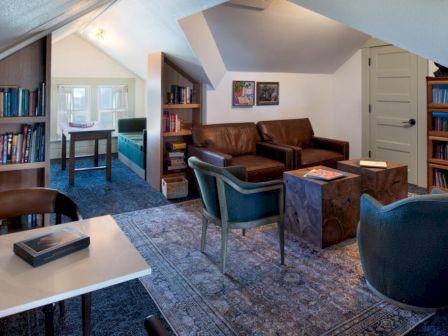 A cozy, book-filled attic lounge with a sofa, armchairs, coffee tables, a patterned rug, and built-in bookshelves lining the walls.